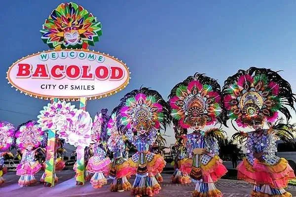 Colorful MassKara dancers in elaborate costumes and smiling masks performing beneath a Welcome Bacolod sign.